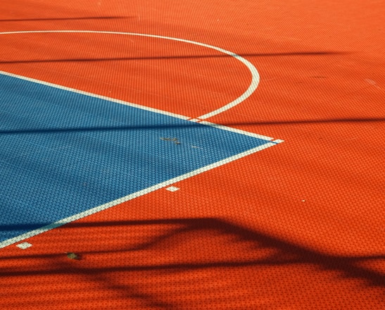 A vibrant sports court with sections of red and blue, featuring crisp white lines marking the boundaries. Shadows from a structure create geometric patterns across the surface, adding depth and interest to the scene.